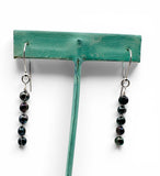 A pair of silver-toned dangle earrings with dark, glossy pearls, displayed on a green T-shaped jewelry stand against a white background.