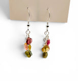 A pair of silver dangle earrings with small polished gemstone charms in pink, yellow, and olive green, displayed on a beige card against a white background.