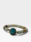 A beaded bracelet featuring natural stone beads in muted beige, brown, and light blue-gray tones, with a prominent oval turquoise-blue stone centerpiece set in a bronze-toned metal frame, shown on a light gray background.