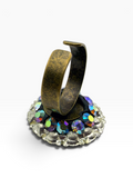 A wide, open cuff ring in an antique bronze finish, displayed on a decorative stand featuring iridescent blue and purple rhinestones on a white background.