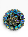 A round vintage-style brooch featuring a cluster of iridescent blue and purple rhinestones set in a gold-tone metal frame, shown against a plain white background.