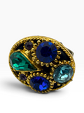 A gold-tone ring featuring a large oval face set with faceted gemstones in deep blue, emerald green, light blue, and small purple accents, photographed on a clean white background.