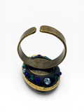 A vintage-style open cuff ring in an antique bronze finish, resting on top of a round black enamel medallion decorated with blue, green, and clear rhinestones, set against a plain white background.