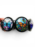 A black metal butterfly body jewelry piece held by a hand against a white background, featuring three circular cabochons with colorful butterfly and floral designs.