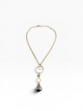 A gold chain necklace featuring a dangle pendant with two stacked gold rings, a small beige bead, and a dark teardrop-shaped stone, displayed on a clean white background.