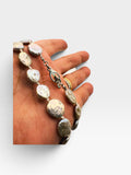 A close-up of a hand holding a beaded necklace with irregular polished stone-like beads and a silver-toned chain clasp, against a plain light background.