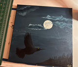 A square canvas painting depicting a black raven in flight under a full moon with clouds, shown resting on a light-colored surface next to a green cutting mat.