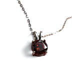 A round faceted dark red gemstone pendant in a silver-toned prong setting on a delicate silver chain, shown against a plain white background.