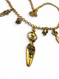 Gold-toned metal charm necklace featuring a central pendant shaped like a stylized female figure with swirling engraved details, surrounded by smaller decorative charms on a white background.