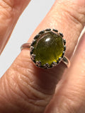 A close-up of a hand wearing a silver ring with a large, oval, olive-green stone.