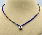 A beaded necklace displayed on a beige jewelry bust. The necklace features a long strand of deep blue beads, a section of small multicolor beads, and a polished silver-toned pendant at the center.