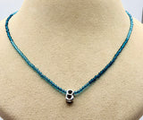 A beaded necklace with small blue beads and a silver letter 'B' pendant, displayed on a beige jewelry bust.