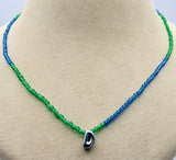 A beaded necklace with a color gradient transitioning from green to blue, featuring a small silver-toned pendant with a dark blue stone, displayed on a beige jewelry bust.