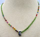 A beaded necklace displayed on a beige jewelry bust. The necklace features a long strand of faceted green beads with a small cluster of multicolor beads near the center, culminating in a polished silver letter 'E' pendant.