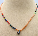 A beaded necklace displayed on a beige jewelry bust. The necklace features a multicolor strand of small round beads on one side and a translucent orange strand on the other, with a polished silver-tone hook-shaped pendant at the center.