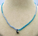 A beaded necklace displayed on a beige jewelry bust. The necklace features faceted beads that transition in color from light icy blue to vibrant turquoise blue, with a small black letter 'L' pendant at the center.