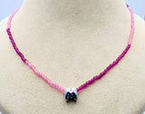 A beaded necklace displayed on a beige jewelry bust. The necklace features small faceted beads that transition in color from light pink at the top to deep magenta/purple at the bottom, with a central silver-toned letter 'M' charm containing dark blue and white enamel.