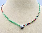 A beaded necklace displayed on a beige jewelry bust. The necklace features a strand of light green faceted beads on one side, a strand of small multicolored beads on the other, and a polished silver letter 'N' pendant at the center.