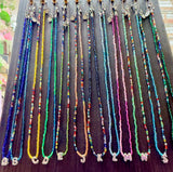 A set of colorful alphabet bead necklaces displayed on a dark wooden hanger. Each necklace features silver letter charms spelling out the alphabet from A to Z, with various vibrant bead strands in blue, yellow, green, pink, orange, and silver.