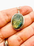 A silver oval pendant with a carved green and blue labradorite iridescent stone, held in a person's hand.