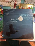 A hand holds a square canvas painting depicting a black bird flying under a full moon in a dark blue night sky with clouds and a tree line. The background shows an art studio desk with paintbrushes, scissors, and supplies.