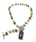 A beaded necklace with a silver-toned toggle clasp and a dangling dog tag charm featuring a paw print, made with mixed earthy and pastel beads on a white background.