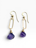 A pair of gold drop earrings featuring faceted purple stone pendants, shown on a clean white background.
