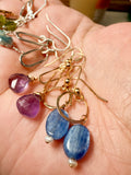 A close-up of handmade dangle earrings held in a person's hand. The earrings feature gold-toned metal hooks and hoops with faceted gemstone drops in purple, blue, and silver, with a small white pearl accent.