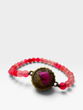 A beaded bracelet featuring translucent pink and red beads with a central oval bronze-toned medallion containing a preserved pink flower, shown on a light gray background.