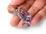 A pair of silver wire-wrapped dangle earrings with faceted purple and lavender gemstones, held in a person's hand against a white background.