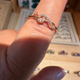 A close-up of a gold ring with a swirled, open design set with small clear stones, worn on a finger. The background shows a jewelry display box with other rings.