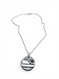 A silver pendant necklace featuring a textured circular disc with a minimalist geometric design and a small red gemstone accent, displayed on a clean white background.
