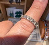 A silver ring with a continuous row of clear rectangular baguette-cut stones, shown worn on a person's finger in a jewelry store setting.