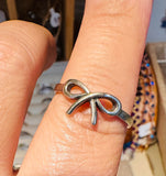 A close-up of a silver bow-shaped ring worn on a person's finger, with a softly blurred jewelry display in the background.