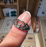 A close-up of a silver ring with a textured, organic knot-like design worn on a finger with a French manicure, set against a blurred jewelry display background.