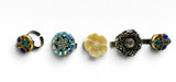 Five vintage-style rings displayed horizontally on a plain white background, featuring various decorative elements including blue rhinestones, a cream flower, and dark filigree.