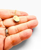 A small oval gold pendant necklace on a delicate gold chain, held in the palm of a hand. The hand also wears a gold ring with dark lettering, against a clean white background.