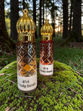Two decorative perfume bottles labeled '#14 Woody Sands' standing on moss in a forest setting. The bottle on the left contains amber liquid and the bottle on the right contains a reddish-brown liquid, both with gold cap details and diamond-patterned glass.