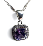 A silver-toned square pendant necklace featuring a cushion-cut purple gemstone surrounded by a halo of small clear stones, suspended from a silver chain on a white background.