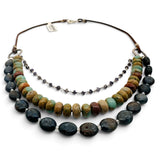 A multi-strand beaded necklace featuring three distinct layers: a long strand of large polished earthy beads, a shorter strand of small dark gray beads, and a brown cord with a silver clasp, displayed on a white background.