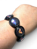 A black bracelet worn on a person's wrist, featuring round glossy charms with colorful cosmic and butterfly illustrations under clear domes.