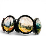 A black cuff bracelet featuring three large oval compartments, each filled with a hand-painted mountain and forest landscape scene.