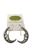 A pair of silver crescent moon earrings with a sleeping face detail, displayed on a white brand card over a gray laptop keyboard.